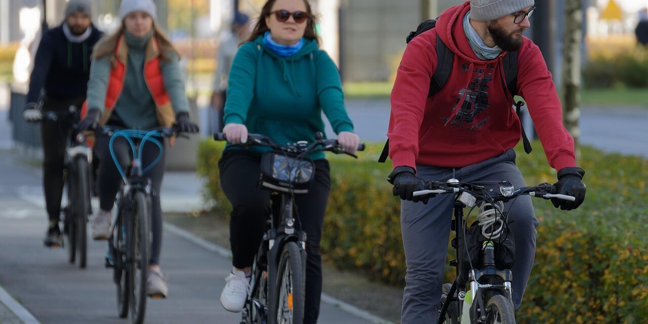Das Bild zeigt eine Gruppe von vier Personen, die auf Fahrrädern auf einem Bürgersteig fahren. Die Personen sind alle warm angezogen, mit Mützen, Handschuhen und Jacken oder Pullovern, was darauf hindeutet, dass es sich um eine kühle Jahreszeit handelt. Der Mann im Vordergrund trägt einen roten Hoodie, eine graue Mütze und eine Brille und hat einen Bart. Die Frau hinter ihm trägt einen türkisfarbenen Hoodie und Sonnenbrillen. Die anderen beiden Personen sind etwas verschwommener im Hintergrund, aber man kann erkennen, dass auch sie warme Kleidung tragen. Der Bürgersteig scheint von Bäumen und Sträuchern gesäumt zu sein, was auf eine städtische oder vorstädtische Umgebung hindeutet.