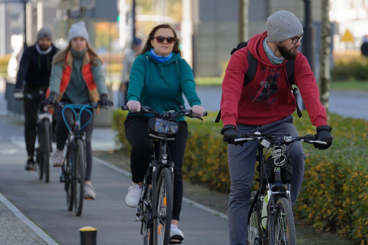 Das Bild zeigt eine Gruppe von vier Personen, die auf Fahrrädern auf einem Bürgersteig fahren. Die Personen sind alle warm angezogen, mit Mützen, Handschuhen und Jacken oder Pullovern, was darauf hindeutet, dass es sich um eine kühle Jahreszeit handelt. Der Mann im Vordergrund trägt einen roten Hoodie, eine graue Mütze und eine Brille und hat einen Bart. Die Frau hinter ihm trägt einen türkisfarbenen Hoodie und Sonnenbrillen. Die anderen beiden Personen sind etwas verschwommener im Hintergrund, aber man kann erkennen, dass auch sie warme Kleidung tragen. Der Bürgersteig scheint von Bäumen und Sträuchern gesäumt zu sein, was auf eine städtische oder vorstädtische Umgebung hindeutet.